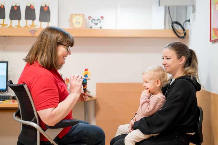 Woman in red shirt plays with toy for child sitting with another woman in a medical office.