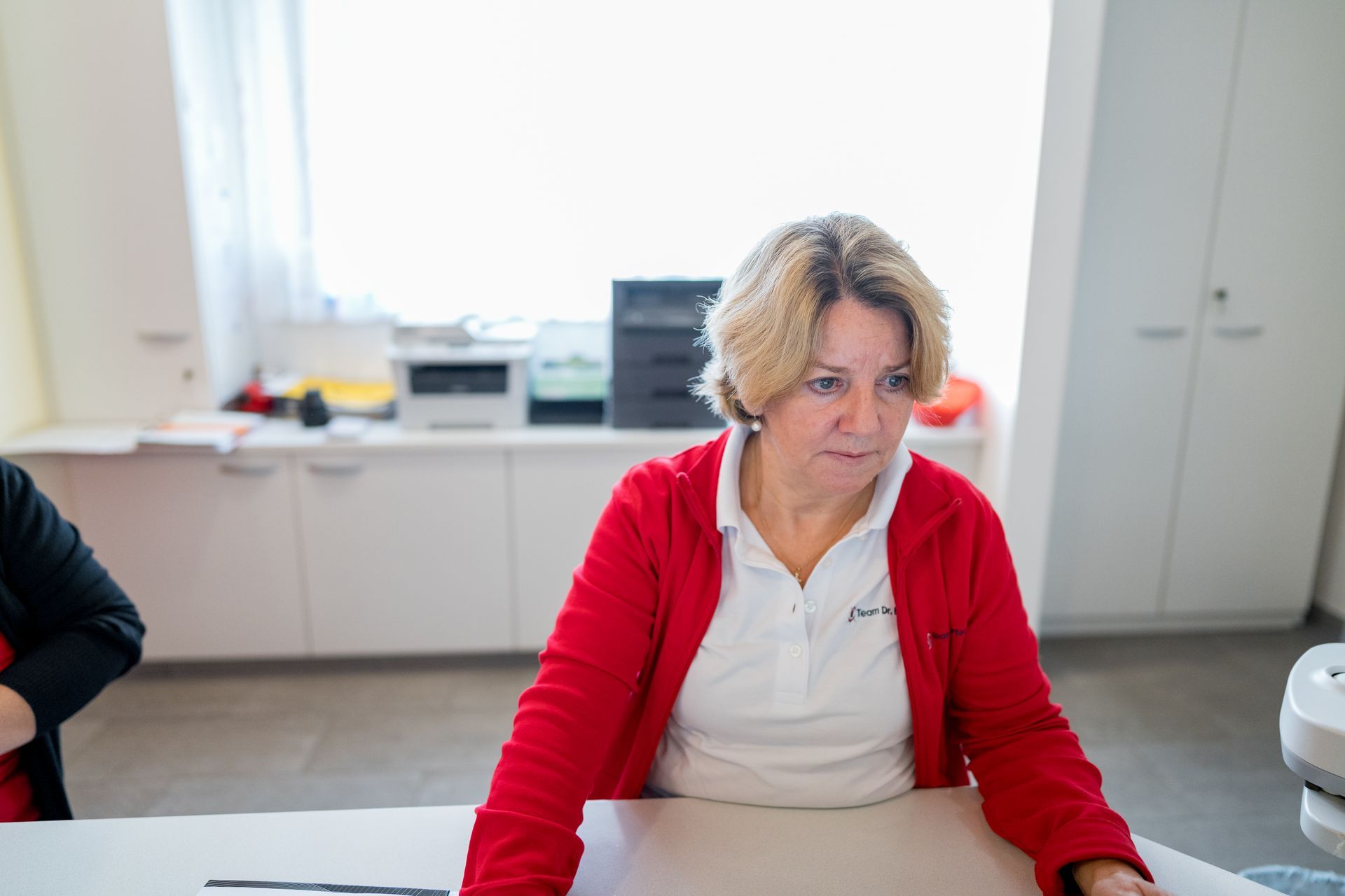 Woman in red jacket looking down at a desk in an office.