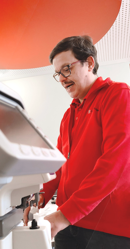 Man in red hoodie, eyeglasses operating a medical device with a touchscreen and control knobs in a well-lit interior.