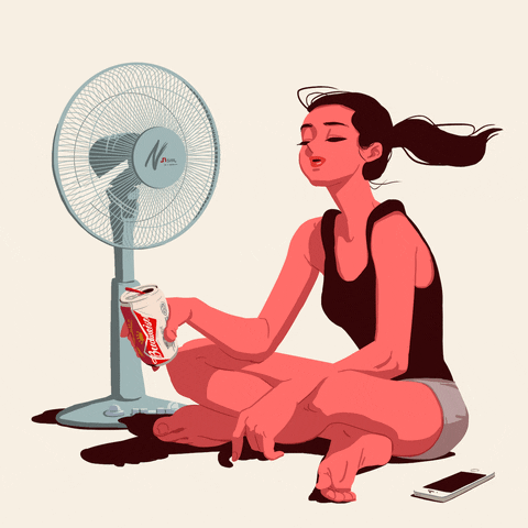 A person in a black tank top sitting cross-legged, holding a can while relaxing in front of a blue electric floor fan.