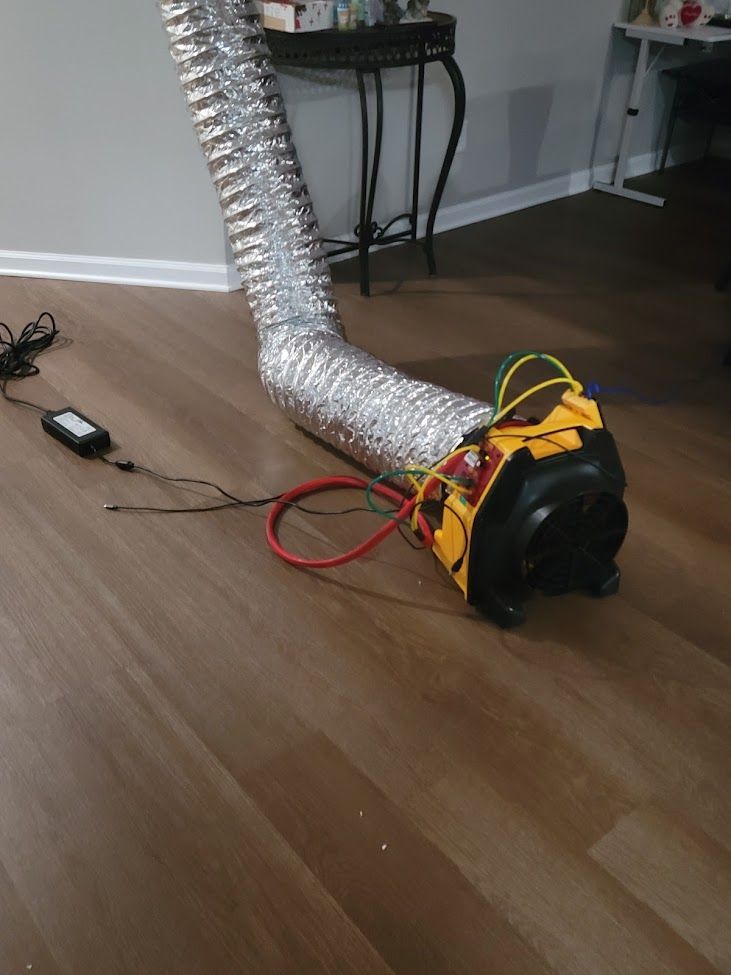 Silver flexible duct attached to a small black-and-orange floor fan on a hardwood floor with cords nearby