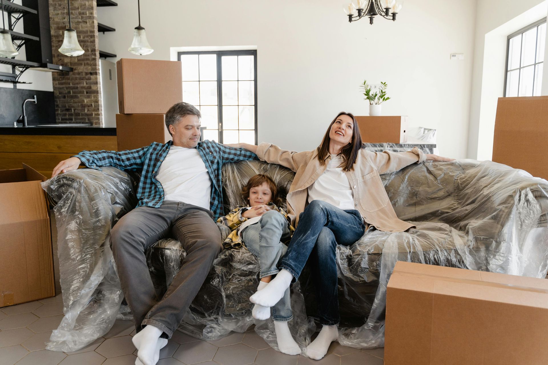 Three people relaxing on a couch in a bright living room with moving boxes around them.
