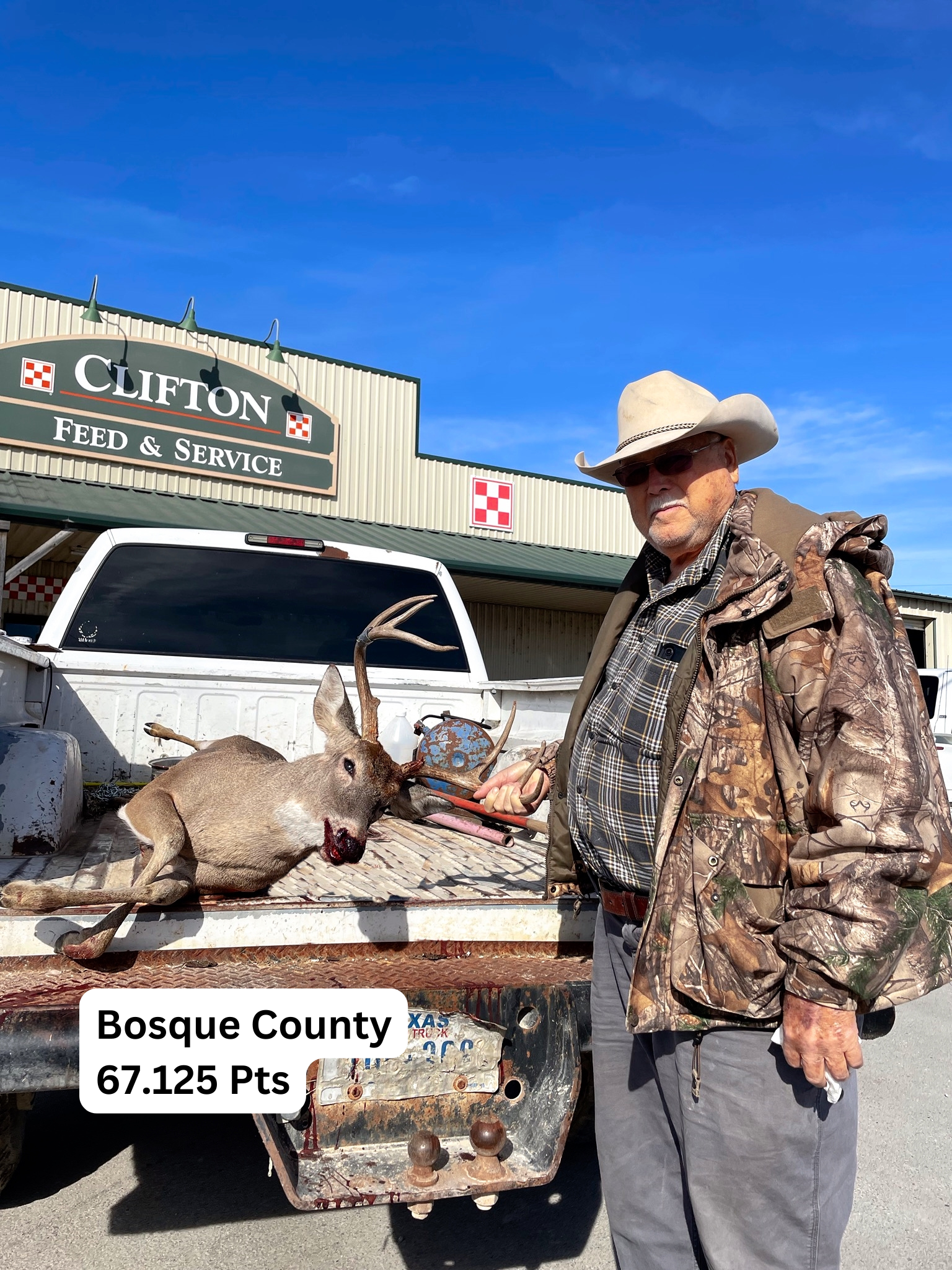 Man in camo next to a deer in a truck bed, in front of a feed store in Basque County.