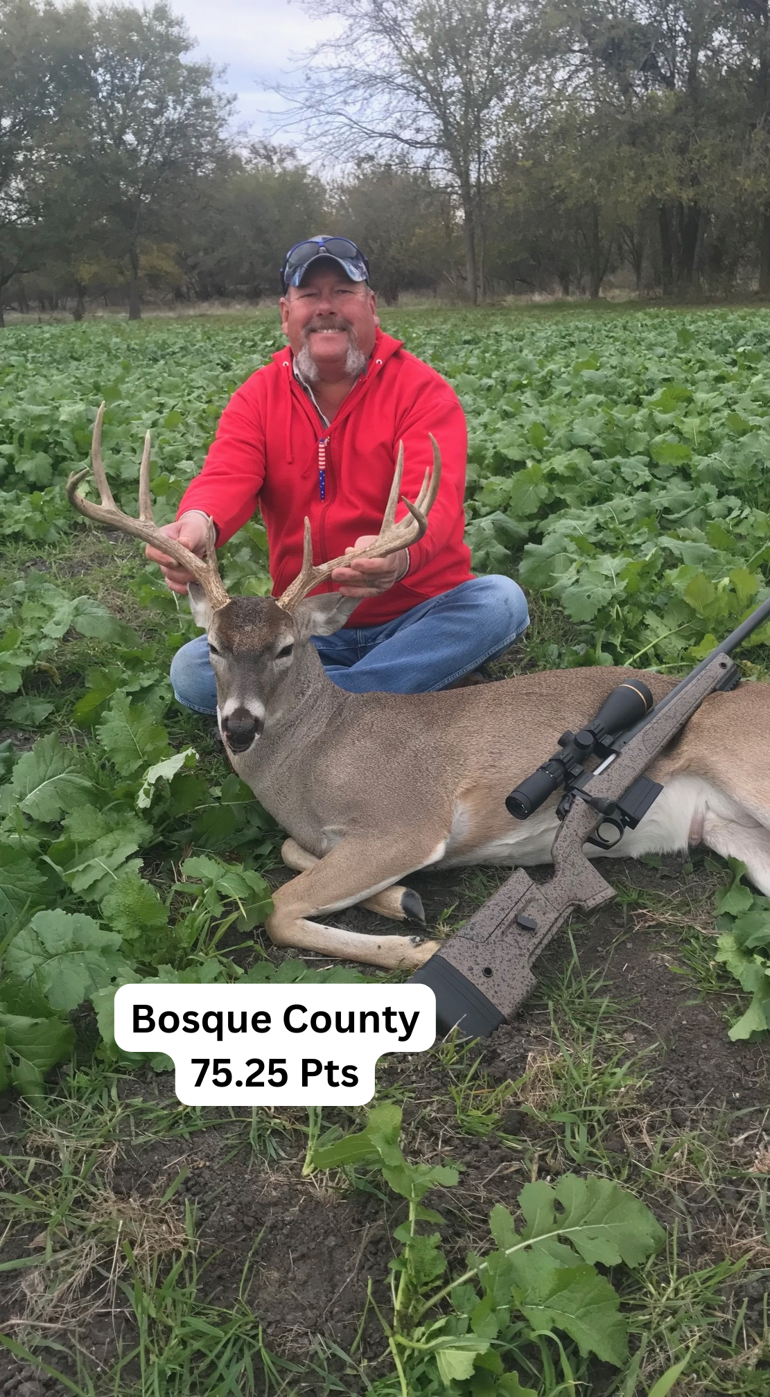 Man with deer, Bosque County. Man smiles, holding antlers. Deer lies on field. Rifle beside deer.