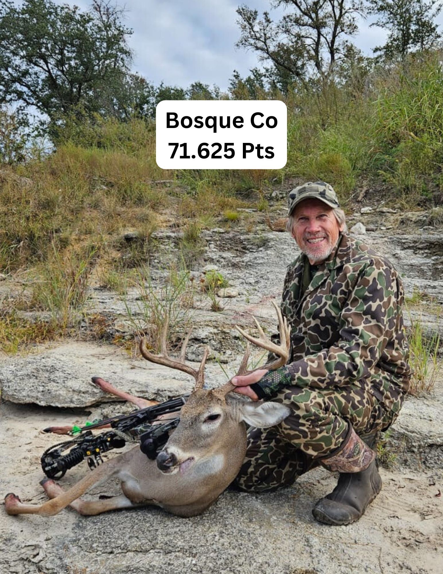 Man kneels with a deer he hunted in Bosque Co, Texas. He wears camo, smiles. Deer has large antlers.