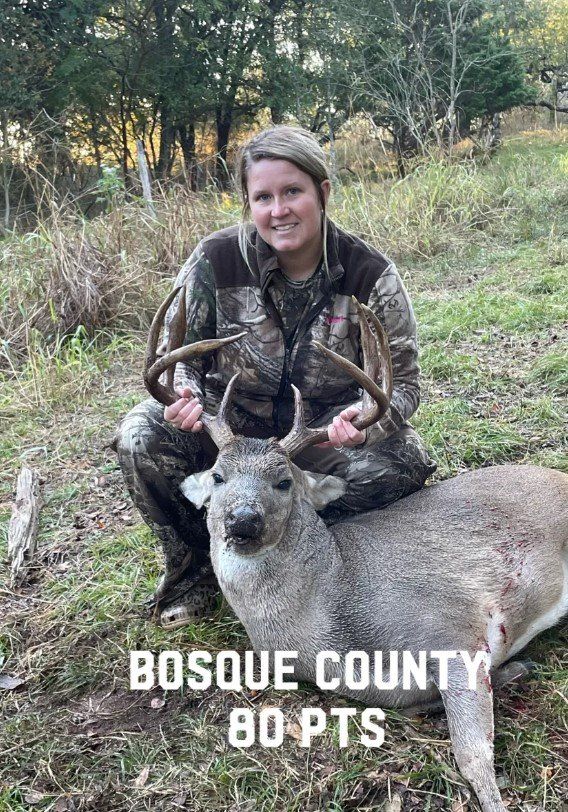 Woman in camo kneels with a large buck in Bosque County, holding its antlers.