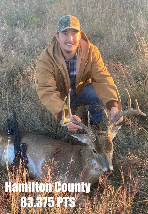 Man kneeling with a large buck in Hamilton County, smile, brown jacket, 83.375 points.