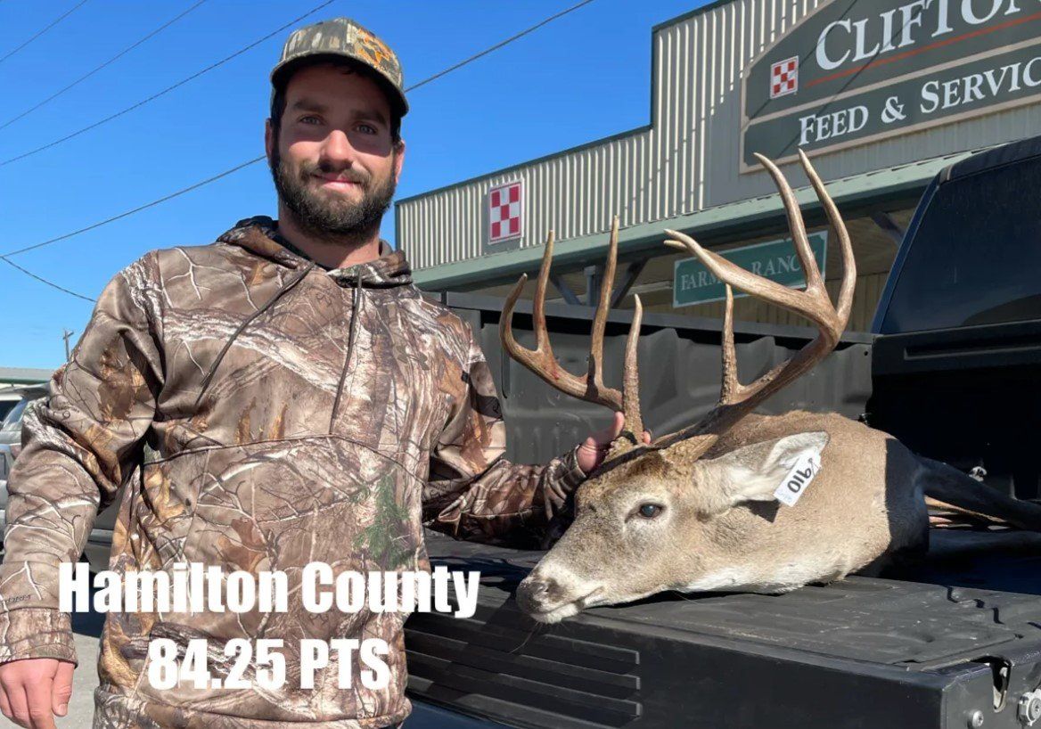 Man in camo with a buck he harvested. Hamilton County, 84.25 PTS. Standing outside a feed store.