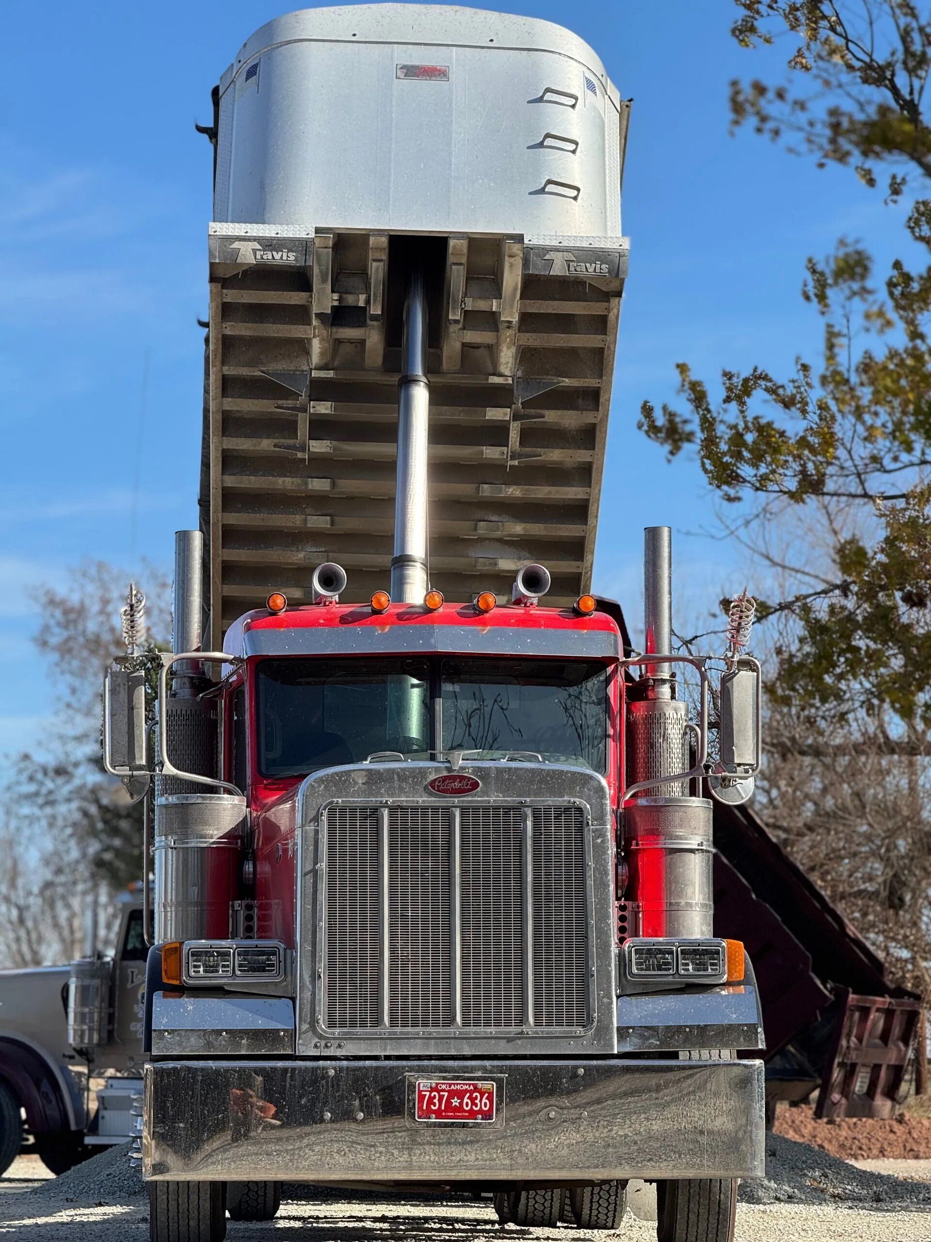 Red semi truck backing under a grain hopper, viewed head-on on a rural dirt road.