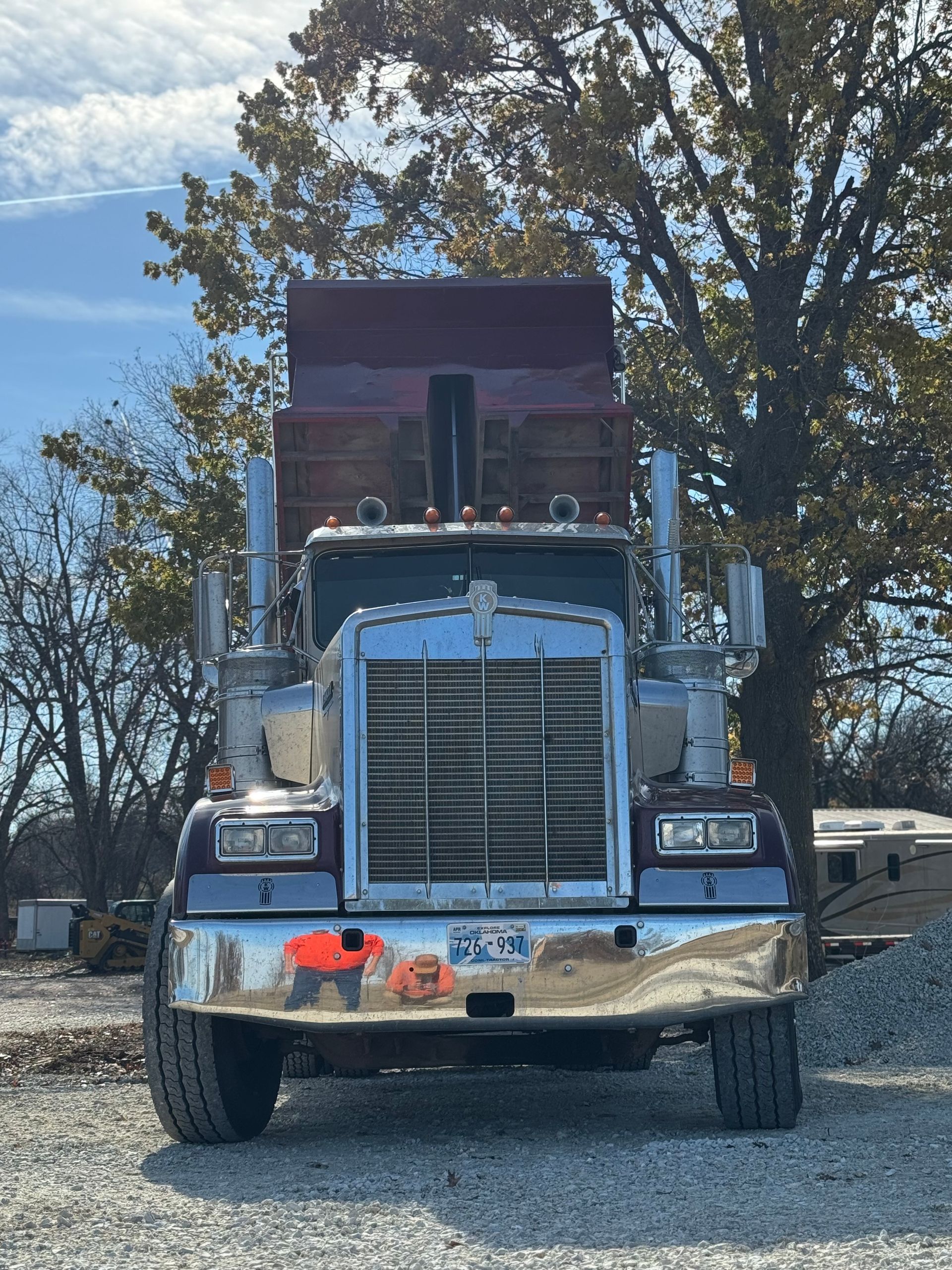 Front view of a blue semi-truck parked on a gravel road with trees in the background.