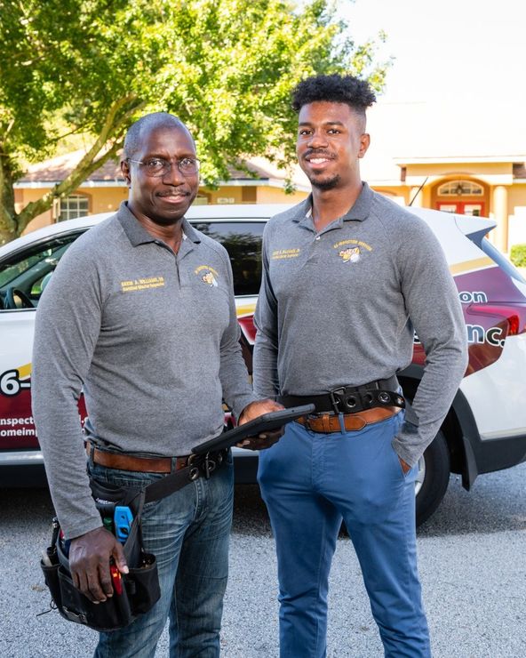 Two professionals in gray shirts and tool belts stand outside, smiling in front of a work vehicle.