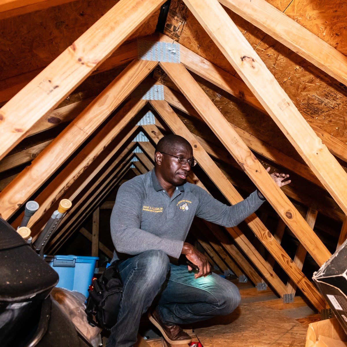 A person inspects wooden roof trusses and structural framing in a dimly lit attic while holding a flashlight.