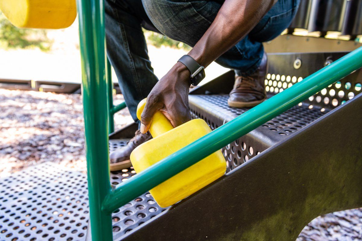 A person holds a yellow plastic component while crouching on a perforated metal playground platform.