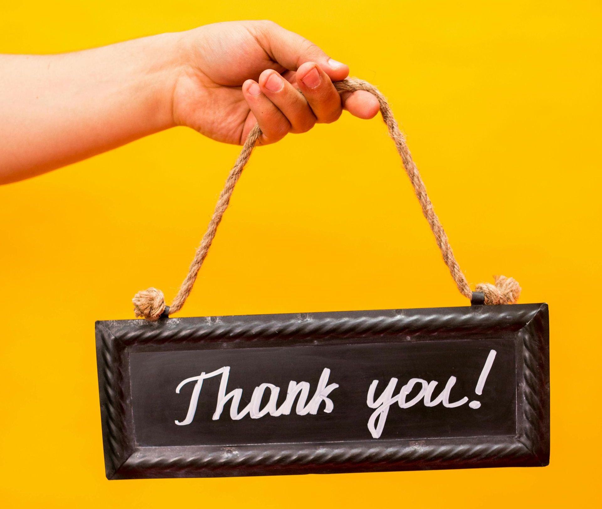 A hand holds a chalkboard sign by a rope handle against a bright yellow background; the sign says