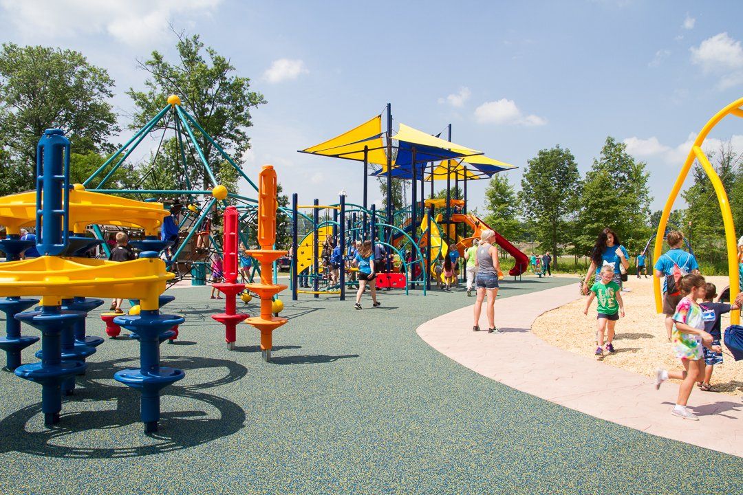 A sunny outdoor playground with colorful play structures, slides, and children playing on a sand pit area.