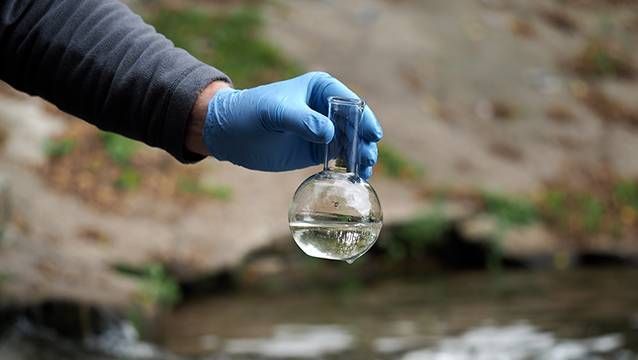 A hand wearing a blue nitrile glove holds a glass flask containing a water sample against an outdoor natural backdrop.