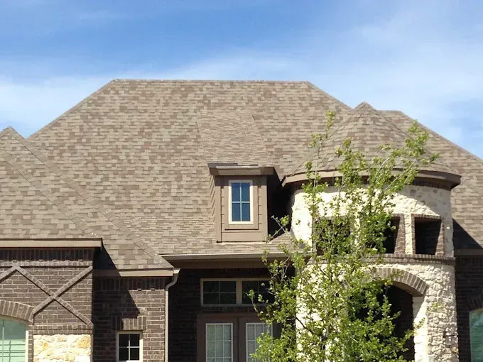 Brown shingled roof of a brick house with a stone turret, under a blue sky.