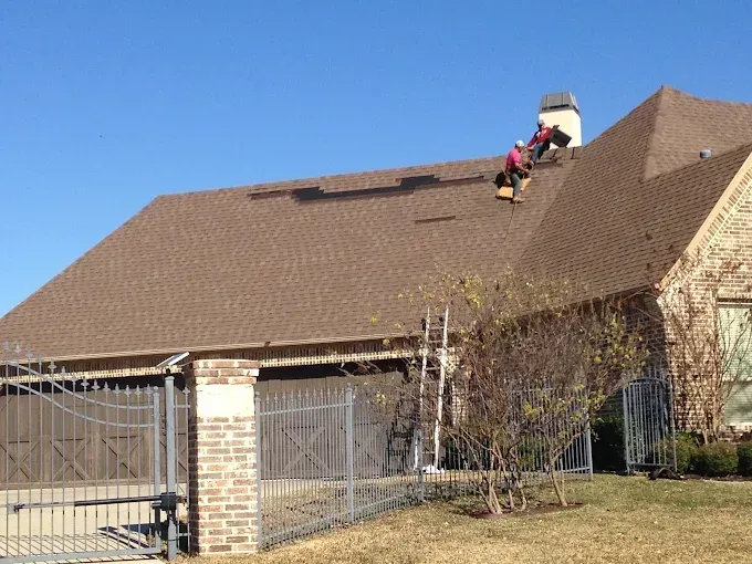 Workers repairing a brown shingle roof on a brick house; clear blue sky.