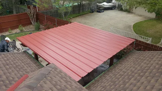 Red metal carport roof over a concrete driveway, with a person working on the structure. Brown shingled roof visible.
