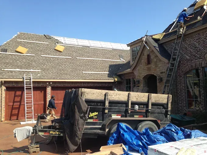 Roofers replacing shingles on a brick house with a trailer and ladders on the ground.