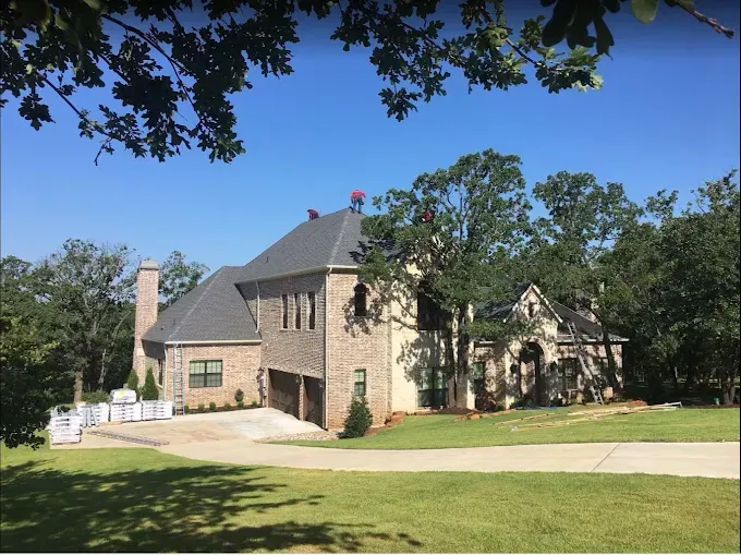 Large brick house with gray roof, surrounded by trees on a sunny day.