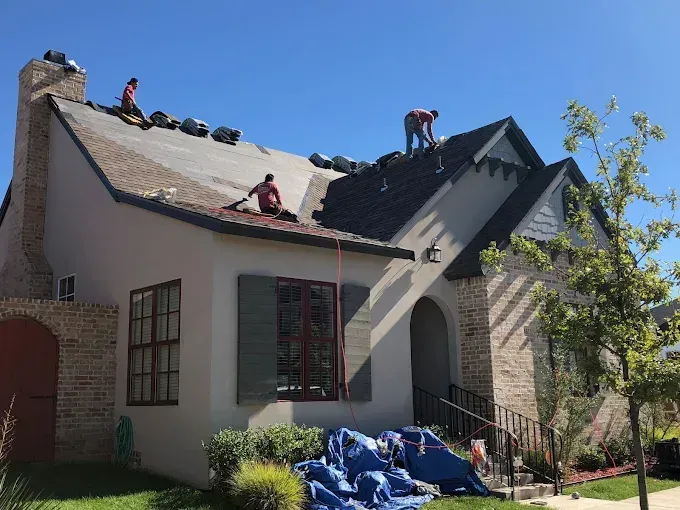 Roofers working on a house roof under a clear blue sky. Blue tarps cover the ground.