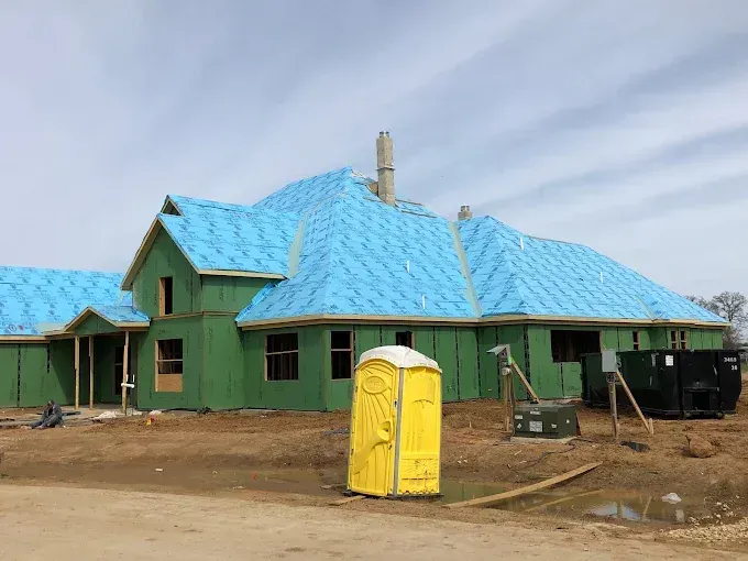 House under construction; blue roof covering, green exterior walls, yellow portable toilet.