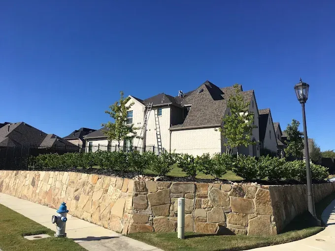 Tan stone wall with hedges, a two-story beige house, and clear blue sky.