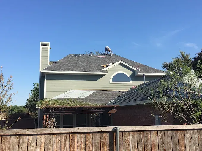 Person on a roof removing shingles, green house with arched window, blue sky, wooden fence.