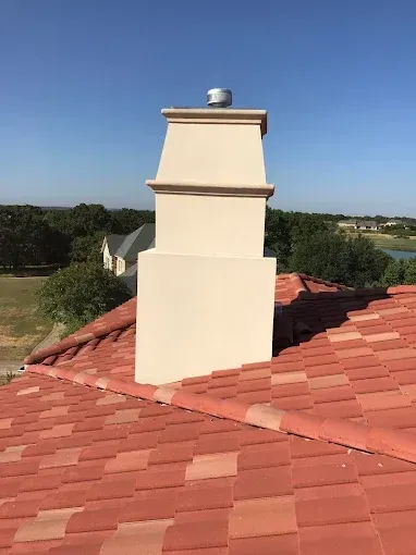 Chimney on a red tile roof, beige stucco, clear sky, with trees and a body of water in the background.