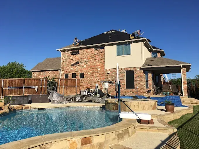 Workers installing roof on a two-story brick home, near a pool, on a sunny day.