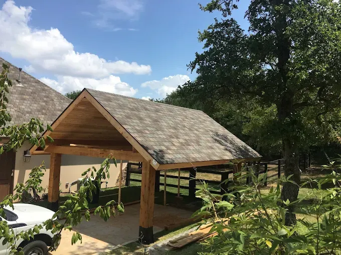 Carport with wood beams, asphalt shingle roof, attached to a tan building on a sunny day.