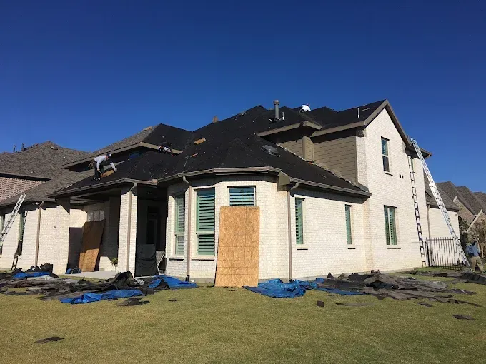 House with roof under construction; workers on roof, ladders, blue tarps, green grass, blue sky.