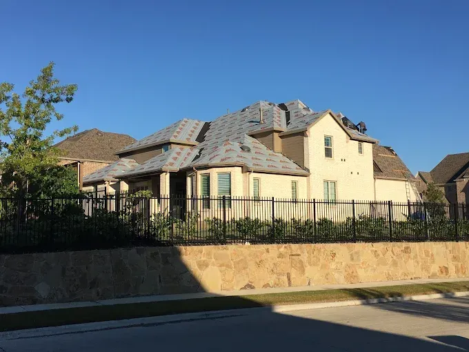 Beige house with a partially shingled roof, black fence, and stone wall, under a blue sky.