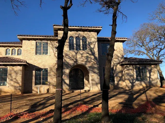 Beige stucco house with arched doorway and multiple windows, trees in foreground, clear blue sky.