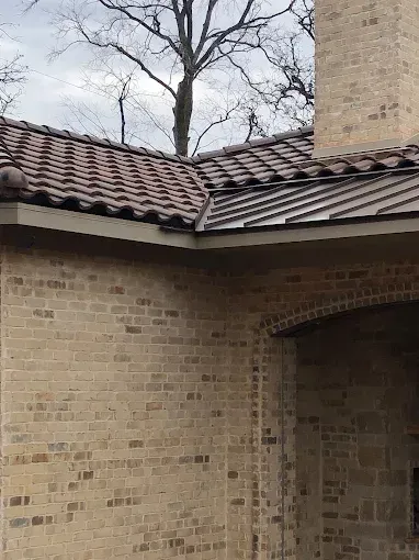 Clay tile roof meets metal roof with brick chimney, building, and trees.