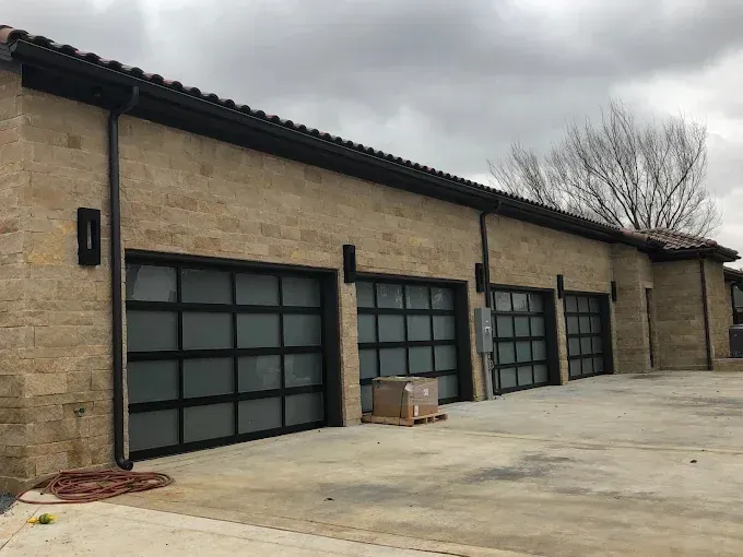 Tan stone building with three garage doors. Black trim and glass panels on the doors. Gray sky.