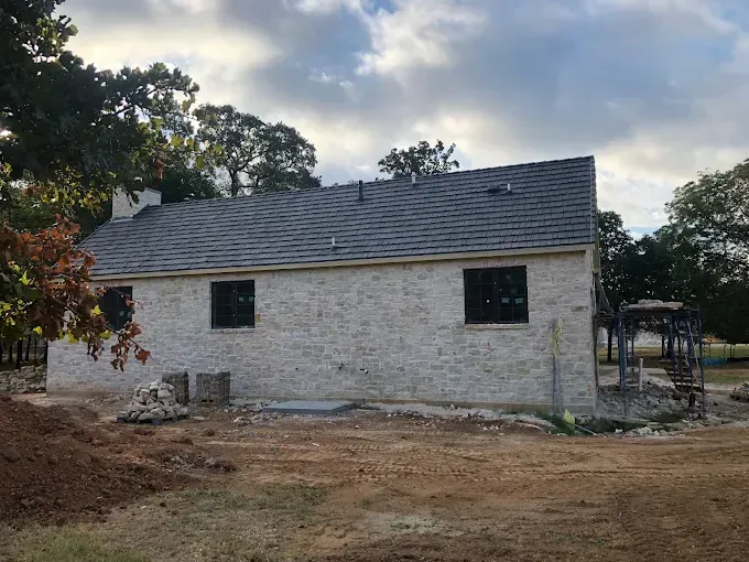 Stone house under construction with dark window frames and gray roof, in a clearing.