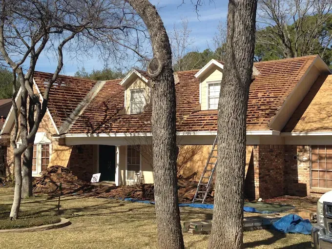 House undergoing roof replacement; brown shingles removed, visible wood, trees in front.