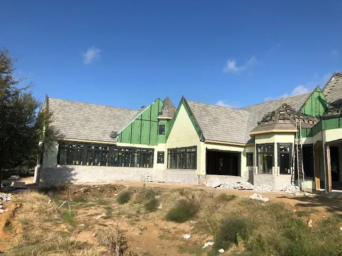 Construction of a large house with gray roof shingles, black window frames, and green siding.