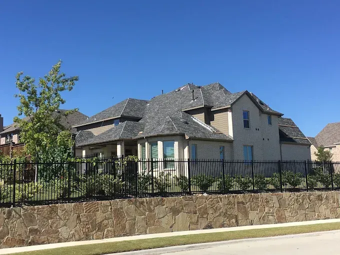 Beige two-story house with gray roof behind a black fence and a stone wall on a sunny day.