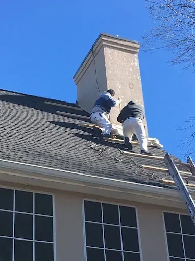 Two workers on a rooftop next to a chimney. One applies something to the chimney. Clear blue sky.