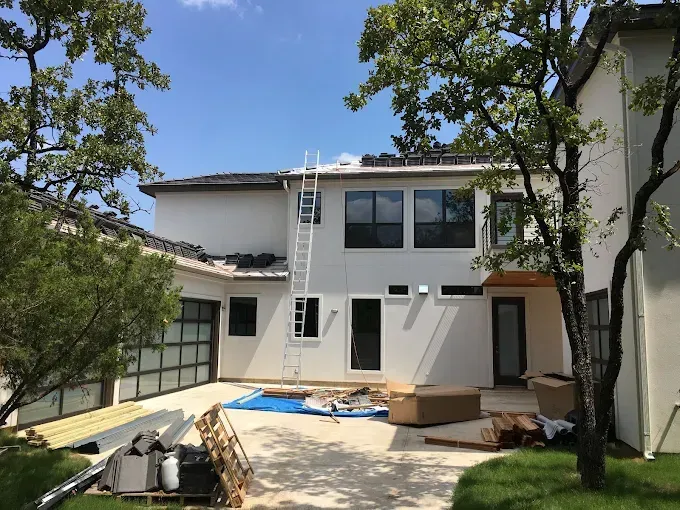 Exterior view of a modern house under construction; ladder on roof, materials on ground, blue tarp, trees, and bright sky.