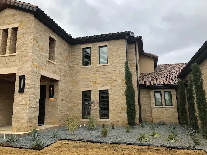 Beige stone house with black-framed windows and a terra-cotta tiled roof under an overcast sky.
