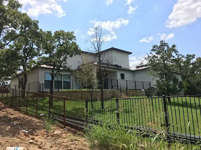 House behind a black fence on a grassy hill; trees frame the background, and blue sky is visible.