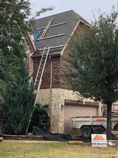 House with partial roof replacement; ladder leans against brick wall; trailer in driveway.