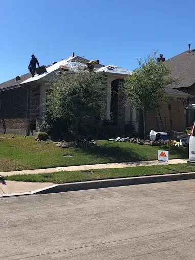 Roofers working on a house under a sunny blue sky. Grass and trees in the front yard.
