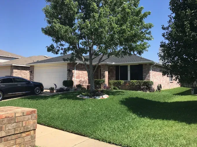 A single-story brick house with a green lawn and a tree in front, next to another house. A car is parked in the driveway.