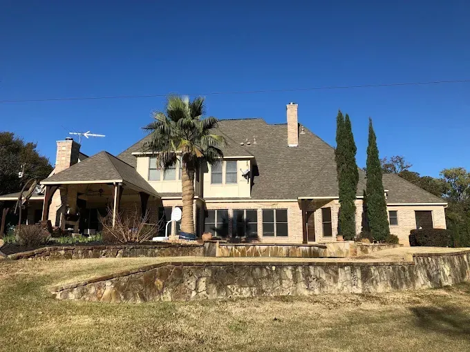 Two-story beige house with a stone retaining wall and palm tree under a blue sky.