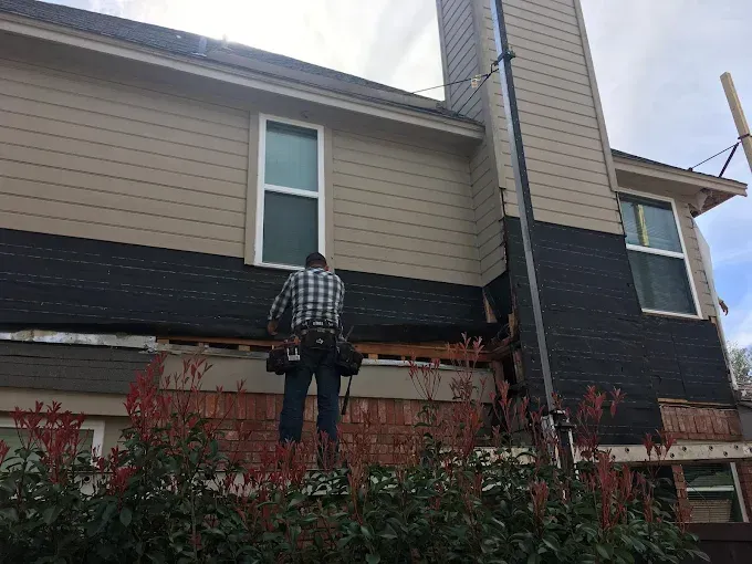 A person on a ladder working on a house with siding. The siding is tan and black.